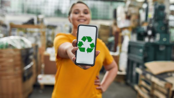 Selective focus on smartphone screen with green recycling sign in hand of happy caucasian young girl working on garbage station, widescreen. Waste sorting and recycling concept